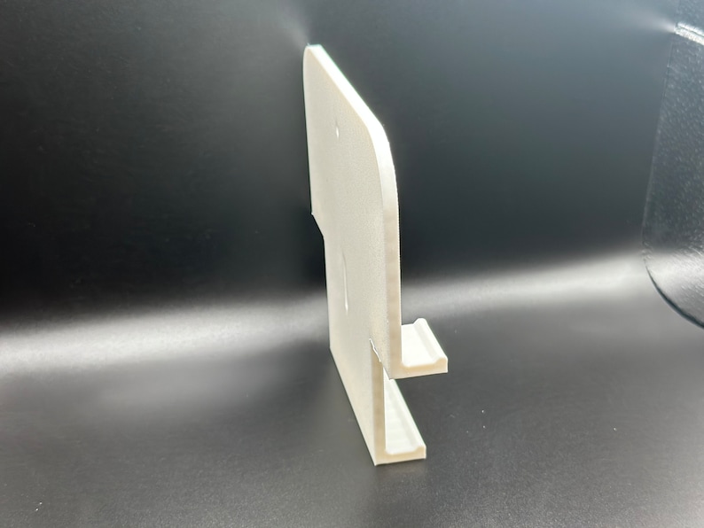 May include: White plastic wall mount phone holder with a single slot for a phone. The holder has a rounded edge and a smooth surface.