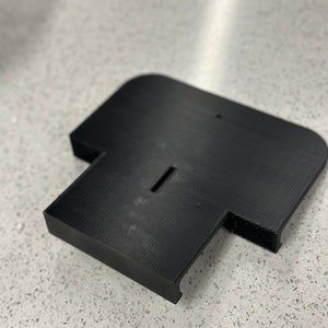 May include: A black plastic base with a slot in the centre and a small raised area on the side. The base is designed to hold a device or object.