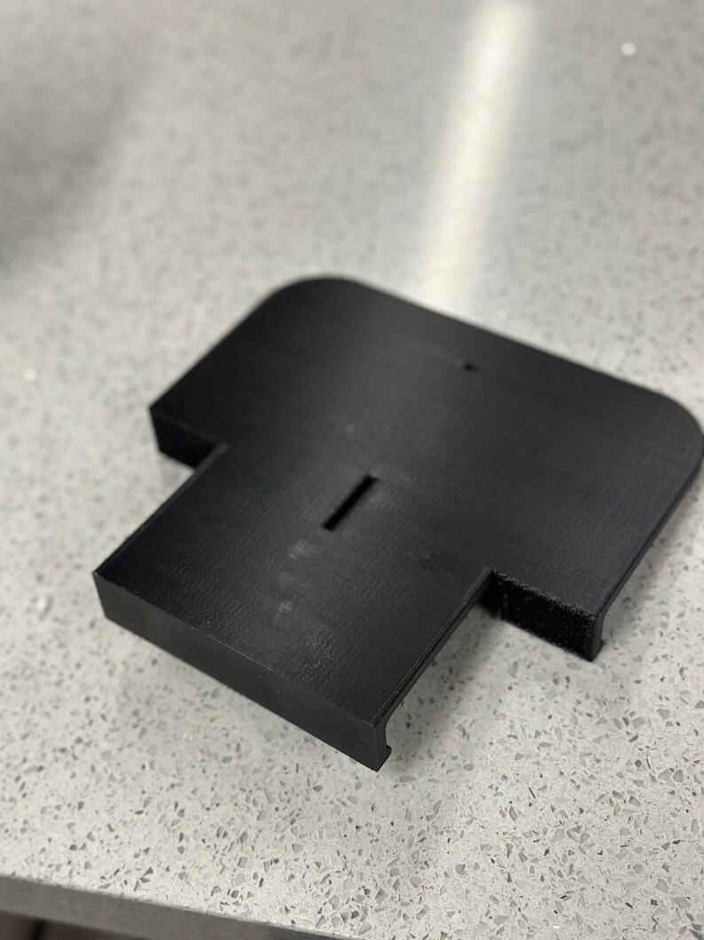 May include: A black plastic base with a slot in the centre and a small raised area on the side. The base is designed to hold a device or object.