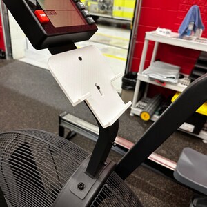 May include: A black and white rowing machine with a white plastic phone holder attached to the arm. The phone holder has a slot for a phone and a small hole for charging cables.