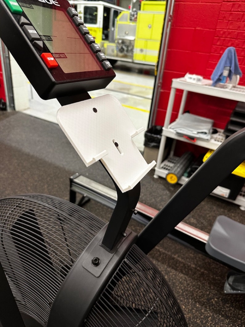 May include: A black and white rowing machine with a white plastic phone holder attached to the arm. The phone holder has a slot for a phone and a small hole for charging cables.