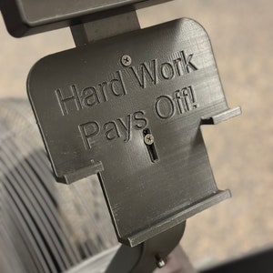 May include: A black 3D printed sign with the text "Hard Work Pays Off!" mounted on a black exercise machine.