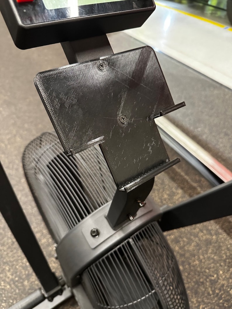 May include: A black 3D printed phone holder attached to a black exercise machine. The holder has a slot for a phone and is mounted on a vertical bar.