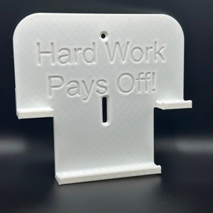 May include: White plastic wall mount with the text "Hard Work Pays Off!" printed on it. The mount has two slots for holding items.