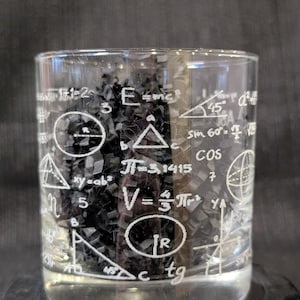 May include: A clear glass tumbler etched with white mathematical formulas and symbols, including equations, geometric shapes, and trigonometric functions. The glass is cylindrical and filled with a dark substance.