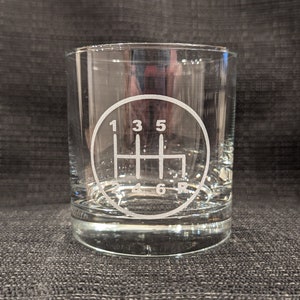 May include: A clear, lowball glass with a white etched design of a car's manual transmission gear shift pattern. The numbers 1, 3, 5, 4, 6, and R are visible. The glass is cylindrical and sits on a dark gray surface.
