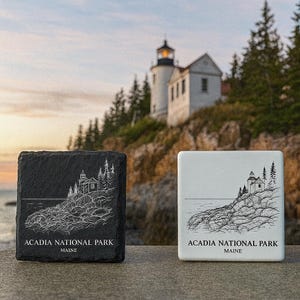 May include: Two square coasters with a lighthouse design. One is black slate, the other white ceramic. Both feature a detailed line drawing of a lighthouse on a rocky coast, with the text "Acadia National Park Maine".