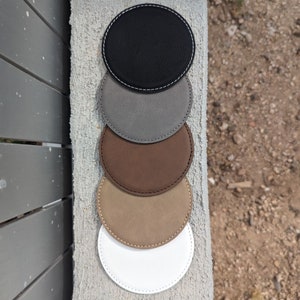 May include: Set of six round leather coasters in various colors, including black, gray, brown, beige, and white. Each coaster has a stitched edge.