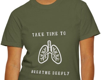 May include: An olive green t-shirt with the text "TAKE TIME TO BREATHE DEEPLY" and a white graphic of lungs. The shirt is made of a soft material and is being worn by a person wearing blue jeans.