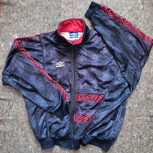 May include: A vintage navy blue Umbro track jacket with a geometric pattern in shades of blue and red accents. The jacket features a full zipper, a red collar, and the Umbro logo on the chest.