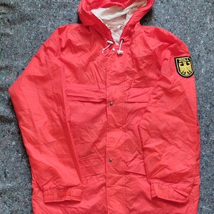 Könnte beinhalten: Eine leuchtend rote Kapuzenjacke mit Reißverschluss und Druckknopfverschluss. Die Jacke hat einen Aufnäher auf dem rechten Ärmel mit dem Wort "ZOLL" und einem deutschen Adlerwappen.