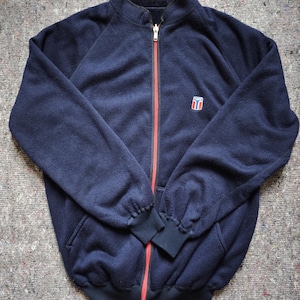 Puede incluir: Chaqueta polar azul marino con cremallera roja y un pequeño emblema en el pecho. La chaqueta tiene cuello alto, mangas largas y puños y dobladillo de canalé negros. Prenda de abrigo informal.