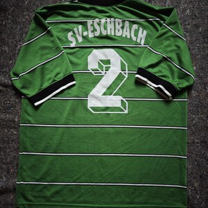 May include: Green and white striped long-sleeved jersey with the text "SV-ESCHBACH" across the shoulders and a large white number "2" on the back. The jersey has black and white trim on the collar and sleeves. The fabric appears to be a lightweight material.