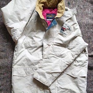May include: A beige Henri Lloyd jacket featuring a tan corduroy collar, a bright pink quilted interior lining, and a patch on the left sleeve that reads THE WORLD RACE and LLOYD with a small flag graphic. The jacket has two front pockets.