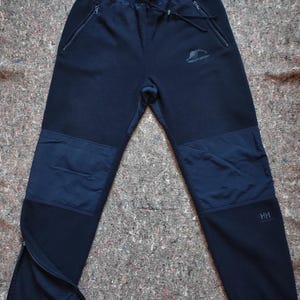 May include: Black fleece sweatpants with a zipper at the bottom of each leg. The sweatpants have a contrasting dark blue panel on each knee and a small logo on the left thigh that reads "Helly Hansen". The text "HH" is printed on the right leg.