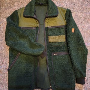 May include: A dark green jacket featuring a textured fleece body and sleeves with smooth olive green shoulder panels. The jacket includes a full-length front zipper, a fold-down collar, two zippered chest pockets, two lower zippered pockets, and black ribbed cuffs.