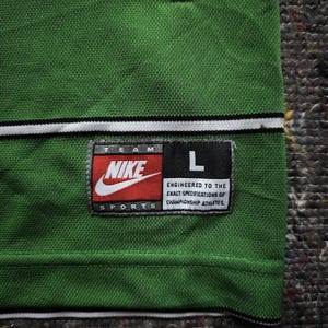 May include: Green athletic jersey with a white and black stripe detail at the neckline. Features a Nike logo patch with the text "TEAM SPORTS" and the size "L". The patch also includes the text "ENGINEERED TO THE EXACT SPECIFICATIONS OF CHAMPIONSHIP ATHLETES."