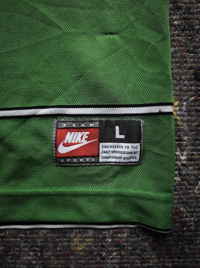 May include: Green athletic jersey with a white and black stripe detail at the neckline. Features a Nike logo patch with the text "TEAM SPORTS" and the size "L". The patch also includes the text "ENGINEERED TO THE EXACT SPECIFICATIONS OF CHAMPIONSHIP ATHLETES."