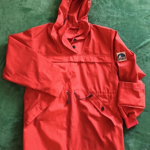 Può includere: Giacca anorak rossa brillante con cappuccio, vita regolabile con coulisse e due tasche anteriori. La giacca presenta una piccola toppa con il logo "Whimbre" sulla manica. Con maniche lunghe e cappuccio.