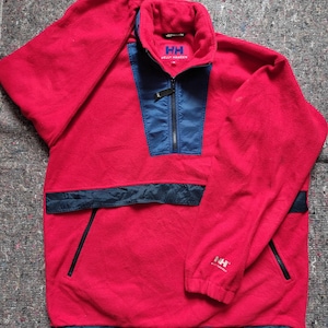 May include: Red fleece pullover with a half-zip closure, blue accents, and the Helly Hansen logo. The pullover has a kangaroo pocket and two side pockets.