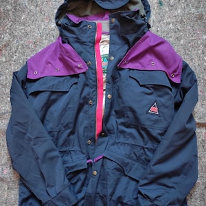 Puede incluir: Una chaqueta azul marino y morada con una cremallera rosa y un logotipo en el lado derecho. La chaqueta tiene una capucha y varios bolsillos.