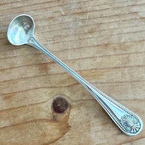 Puede incluir: Una cuchara de sal de plata con un mango con cuentas y un extremo decorativo. La cuchara tiene un cuenco redondo y un mango largo y delgado.