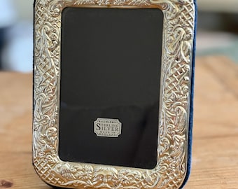 May include: A rectangular silver picture frame with ornate scrollwork and a dark blue velvet backing. The frame has a small oval accent at the top and a label that reads "Hallmarked Sterling Silver Made in England".