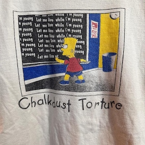 May include: A white t-shirt featuring a cartoon image of a yellow character writing on a blue chalkboard. The chalkboard displays the text "Let me live while I'm young" repeated several times. The text "Chalkdust Torture" is written beneath the image.