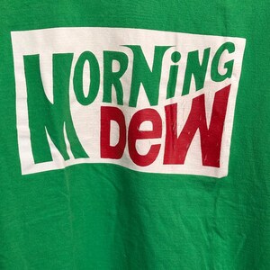 May include: Green crew neck t-shirt with the words "MORNING DEW" printed across the front in green and red lettering on a white rectangular background. The shirt is made of a soft material.