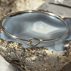 May include: A silver bangle bracelet with a heart-shaped clasp. The bracelet is resting on a gray and white agate stone.