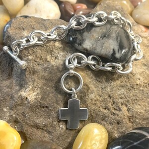 May include: A silver chain bracelet with a silver cross charm. The bracelet is on a bed of smooth stones.