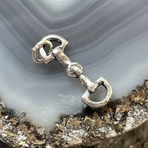 May include: A silver charm in the shape of a horse bit. The charm is detailed and has a rustic finish.