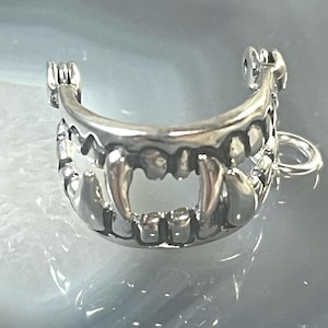 May include: Silver charm in the shape of a set of teeth with fangs. The charm has a loop for attaching to a bracelet or necklace.
