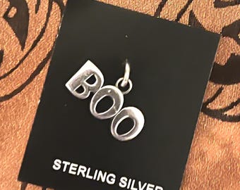 May include: A sterling silver charm spelling "BOO" for Halloween. The charm is attached to a black tag that reads "STERLING SILVER". The background is a patterned orange and brown fabric.