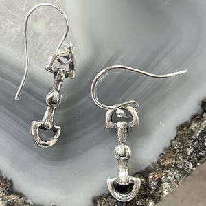 May include: A pair of silver-toned earrings in the shape of a horse bit. The earrings have a hook closure and are displayed on a gray and white marbled surface. The earrings are detailed with a rounded top, a central bar, and a curved bottom.