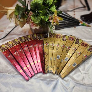 May include: A collection of henna cones in red and gold packaging, arranged in a fan shape. The boxes feature floral and peacock designs, with the brand name "Kashee's" visible. A vase of flowers is in the background.