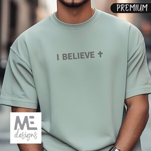 May include: A light green short-sleeved t-shirt with the text "I BELIEVE +" printed across the chest. The shirt has a crew neck and a relaxed fit. The word "PREMIUM" is in the upper right corner. The logo "ME designs" is in the lower left corner.