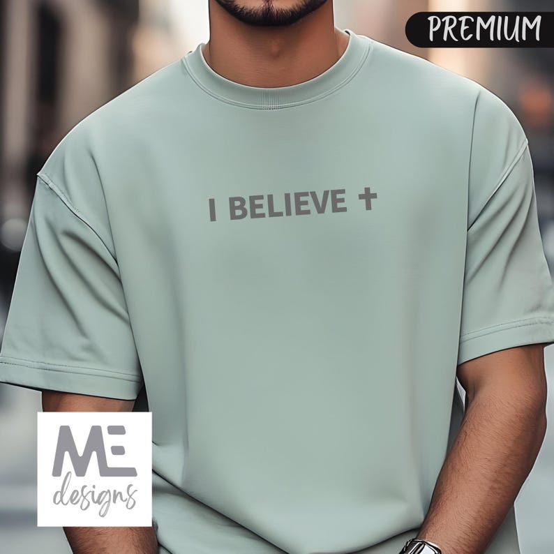 May include: A light green short-sleeved t-shirt with the text "I BELIEVE +" printed across the chest. The shirt has a crew neck and a relaxed fit. The word "PREMIUM" is in the upper right corner. The logo "ME designs" is in the lower left corner.