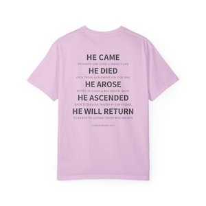 May include: A light pink t-shirt with the text "HE CAME...HE WILL RETURN" in gray. The text is a religious message from 1 Corinthians 15:3-7. The shirt is a short-sleeved crew neck.