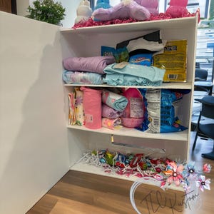 May include: A white shelf unit filled with various items. The top shelf displays plush bunny figures and decorative items. The middle shelves hold folded clothing, snacks, and other packaged goods. The bottom shelf contains candy and decorative filler.