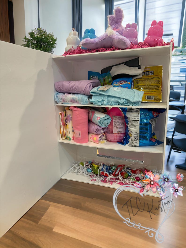 May include: A white shelf unit filled with various items. The top shelf displays plush bunny figures and decorative items. The middle shelves hold folded clothing, snacks, and other packaged goods. The bottom shelf contains candy and decorative filler.