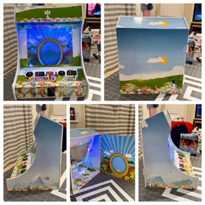 May include: A custom-made arcade machine with a vibrant, colorful design. The front features a screen with a blue and green graphic, and a control panel with buttons and joysticks. The sides and back are decorated with a landscape scene, including a blue sky, clouds, and a rainbow.
