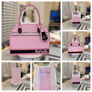 May include: A pink paper handbag with white handles and black accents. The bag features a black stripe and the name "Sarah" in silver script. A black tag with a gold emblem hangs from the handle. The bag is displayed on a white rotating surface.