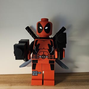 Op de afbeelding: Een rood en zwart Lego minifiguur van Deadpool met een zwart pistool en twee zilveren zwaarden.