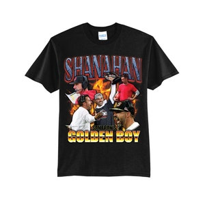 May include: Black t-shirt featuring a collage of images and the name "SHANAHAN" in a retro font. Below the name, the text "SAN FRAN'S GOLDEN BOY" is displayed in a gold and orange font. The shirt has a crew neck and short sleeves.