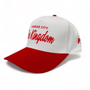 May include: White baseball cap with a red brim. The front of the cap has the text "Kansas City The Kingdom" in red cursive lettering.
