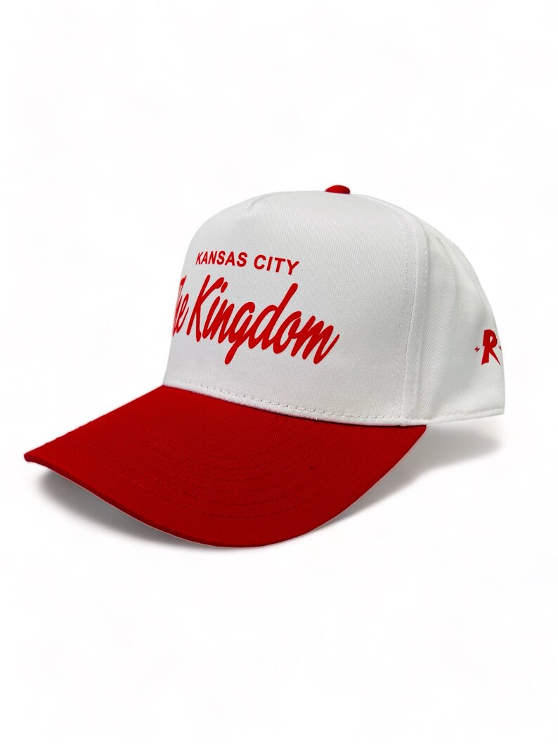 May include: White baseball cap with a red brim. The front of the cap has the text "Kansas City The Kingdom" in red cursive lettering.