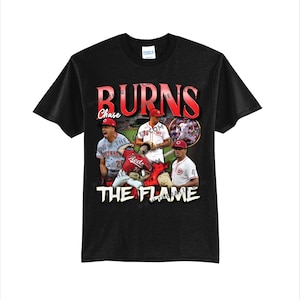 May include: A black short-sleeve t-shirt featuring a graphic print of baseball player Corbin Burnes in various game poses. The text BURNS appears in large red letters at the top, with THE FLAME in white at the bottom and Chase in script.