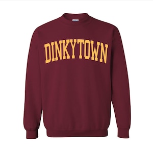May include: A maroon sweatshirt with the word "DINKYTOWN" printed in gold block letters across the chest. The sweatshirt has a crew neck and long sleeves. The background is plain white.