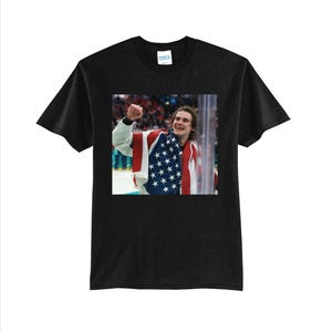 May include: Black t-shirt featuring a photograph of a person wearing a jacket with the American flag. The person is smiling and raising their fist in the air. The background is blurred, suggesting a sporting event.